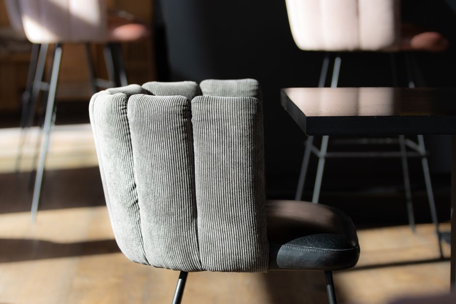 A close-up of a modern office chair with a grey, corduroy upholstered backrest and black padded seat, positioned at a dark wooden table in a well-lit office space. The surface of the table is clean an